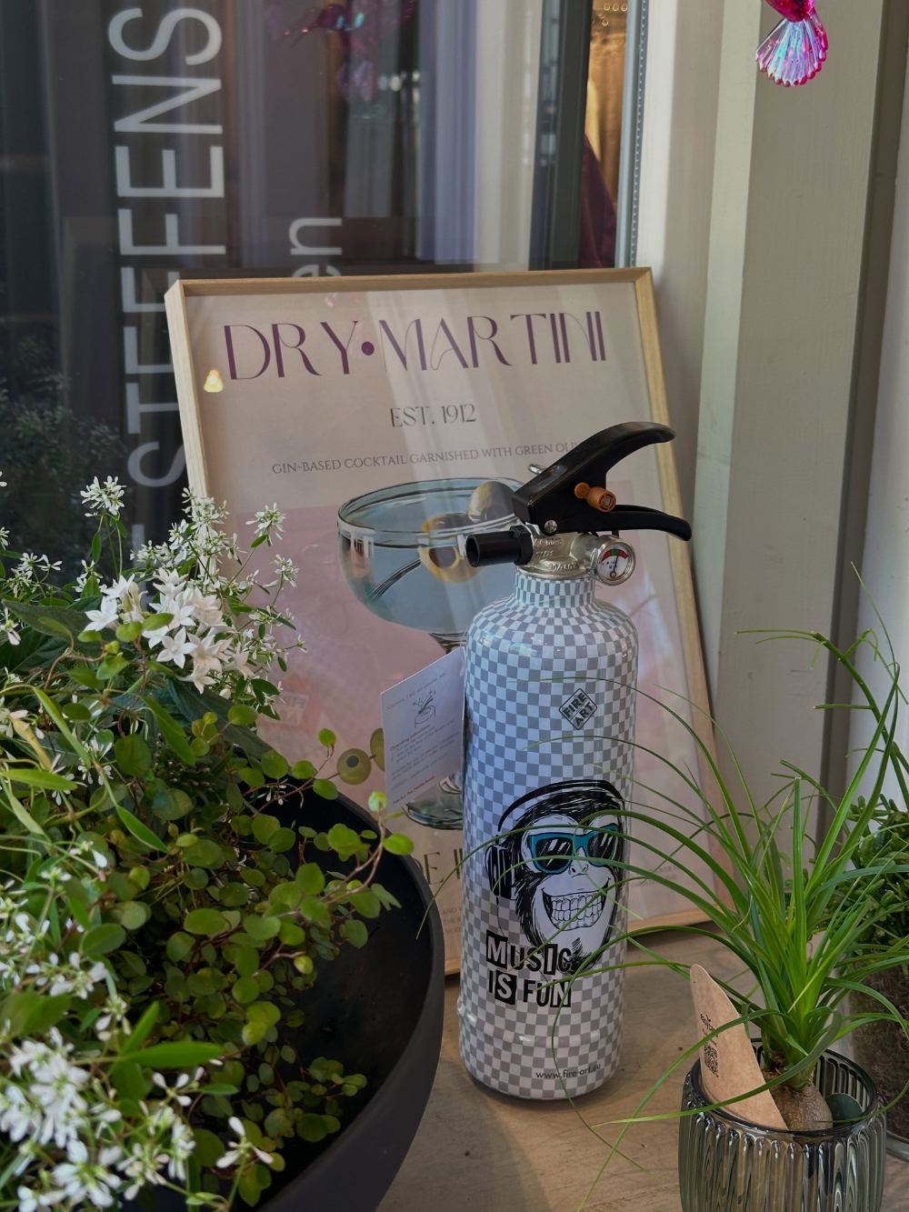 Geschenkideen bei ELLA: bedruckter Design-Feuerlöscher vor einem Dry-Martini-Poster, umgeben von Pflanzen