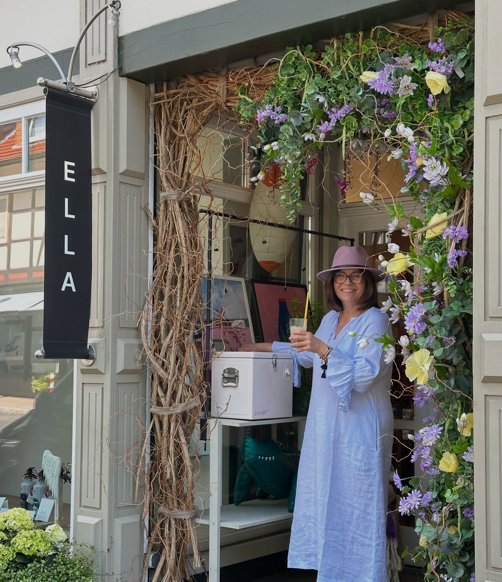 Ella im hellblauen Leinenkleid im blumengeschmückten Eingang ihres Concept Stores in Dransfeld