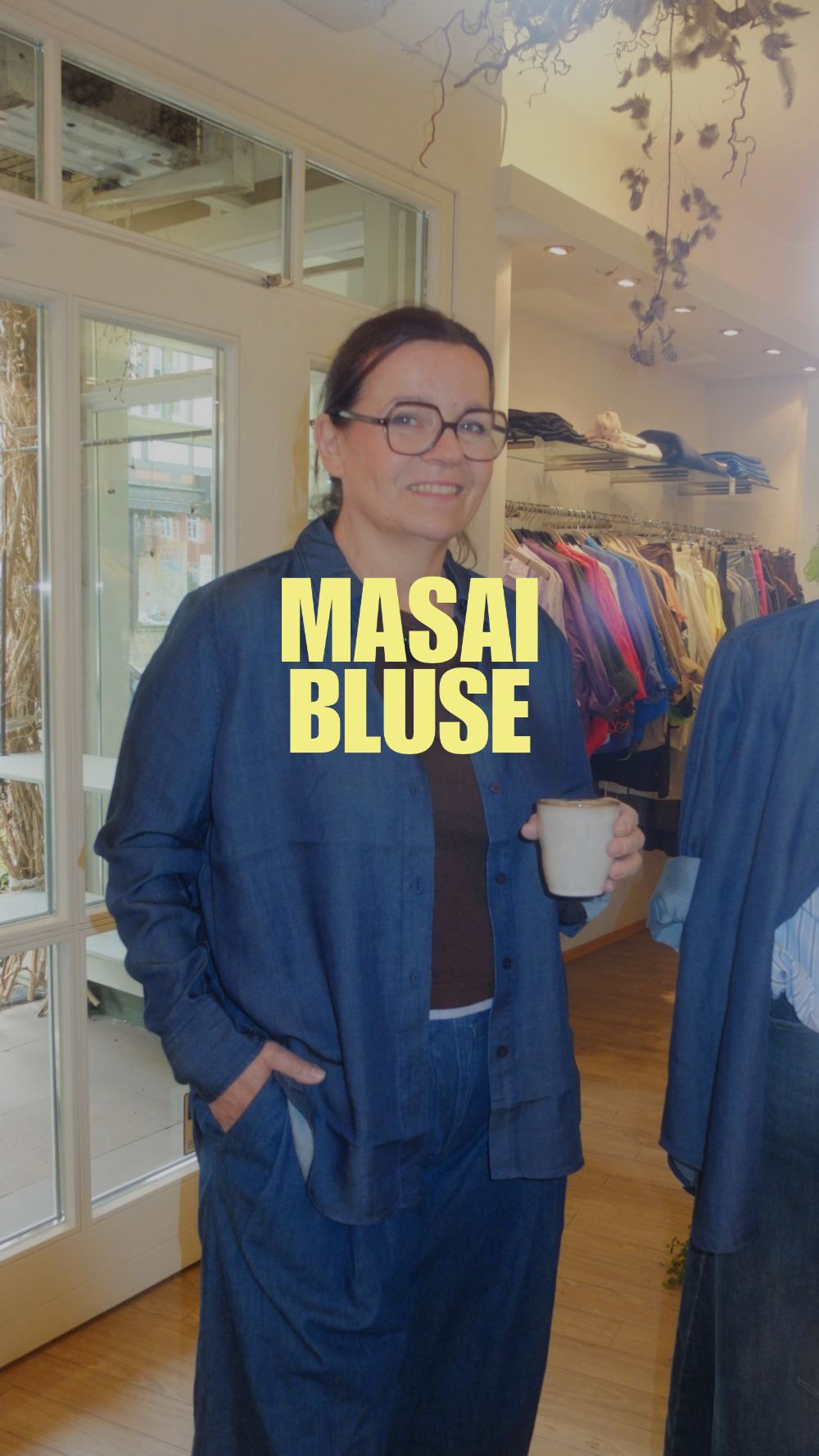 Ella im Laden in einer weiten Jeansbluse von Masai, mit Kaffeebecher in der Hand